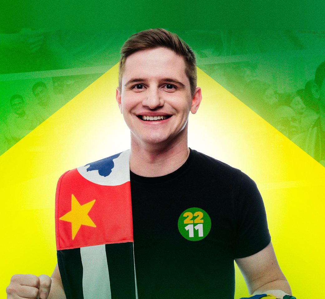 Lucas Pavanato sorrindo, com a bandeira de São Paulo no ombro e o adesivo 2211 na camiseta